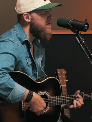 A man with a beard wearing a denim jacket and a baseball cap is playing an acoustic guitar and singing into a microphone. The image is a medium shot, focusing on the man's upper body and the guitar. The lighting seems to be from an indoor environment.