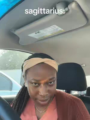 A selfie taken from inside a car. The main subject is a woman with a neutral expression, wearing a headband. The image has a slight angle, and the car's interior, including the sun visor, is visible in the shot. The background includes parts of the outside environment, providing context for the location.