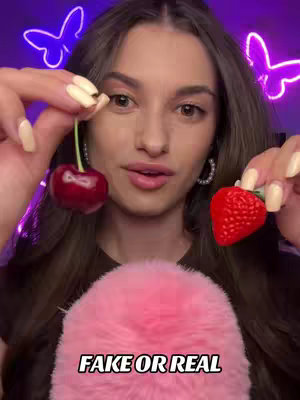 A woman holds a cherry and a strawberry, posing for the camera. The background features neon butterfly lights, and a pink fluffy ball in the foreground. The text "FAKE OR REAL" appears below the woman.