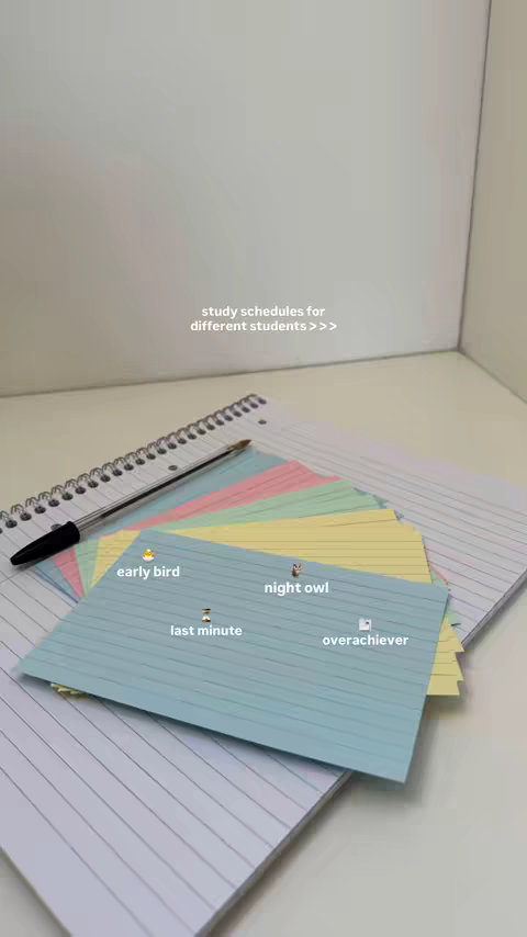 A top-down view of a desk with a white background, featuring a spiral notebook, a black pen, and a colorful stack of index cards. The cards are arranged in a fan shape, with each card labeled and decorated with emojis representing different study styles: an early bird (chicken emoji), a night owl (owl emoji), the last minute (hourglass emoji), and an overachiever (trophy emoji). The text "study schedules for different students >>>" is printed above the notebook cards. The image's composition is clean and organized, conveying a study and organizational theme.
