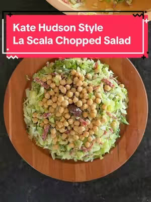A top-down shot of a chopped salad on a wooden plate, set against a dark background. The salad is predominantly green, with a base of chopped lettuce. The salad contains marinated chickpeas, salami, dressing, cucumbers, and fresh basil. The plate is circular and placed centrally, with the salad taking up most of the frame.