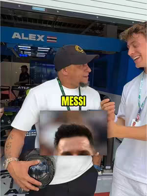 A selfie featuring two men in a garage setting. The man on the left is older, wearing a black cap and white shirt, holding what appears to be a soccer ball. The man on the right, younger, smiles. Between them is a graphic displaying a close-up of Lionel Messi's face, with a yellow text overlay that reads 'MESSI'. The background includes machinery and logos of a racing team.