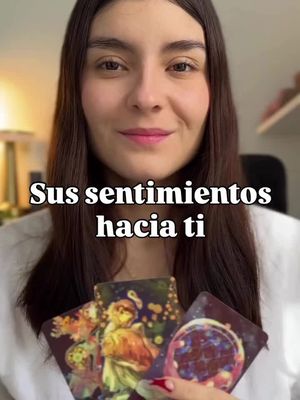 A woman with long dark hair is centered in the frame, smiling slightly and holding three tarot cards. Above her is a text overlay that says "Sus sentimientos hacia ti" (Your feelings towards you) in a white, bold font. The woman's eyes, lips, and overall expression project a sense of warmth and openness. The background is softly blurred, drawing focus to the woman and the cards. The overall color palette is warm with a focus on natural light and a hint of orange tones, indicating her connection to the content.