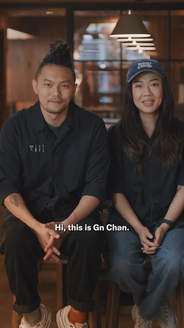 Two people, one male and one female, are seated in front of a blurred background, likely indoors. The man on the left has dark hair styled up, a dark shirt and corduroy pants, and white sneakers with orange socks. He has a serious expression. The woman on the right is wearing a cap, jeans and a black shirt, smiling. The man's elbows rest on his knees with his hands clasped. A text overlay says 'Hi, this is Gn Chan.'