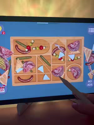 The cover image displays a screen from a mobile game. The game appears to involve arranging food items such as sausages, tomatoes, and cheese on a wooden tray puzzle. A hand with the stylus is adjusting pieces.