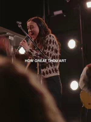 A woman with long brown hair is singing into a microphone on a stage. She is wearing a patterned sweater and black pants. The background is dark, with stage lights visible. Text overlay 'HOW GREAT THOU ART' is at the bottom.