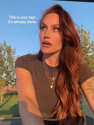 A woman with long auburn hair is looking away from the camera with a thoughtful expression. The background shows a clear blue sky and some foliage. The woman is wearing a dark t-shirt and layered necklaces.