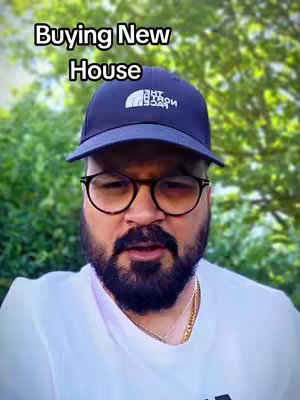 A man wearing a navy blue baseball cap, round eyeglasses, and a white t-shirt against a blurred green background. The overlay text reads 'Buying New House'.