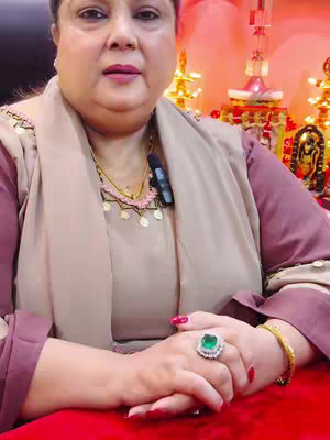 A woman with light skin and short, dark hair poses in front of a decorative background. She wears a maroon top and a cream scarf with a small microphone clipped to her shirt. Her jewelry includes a necklace, bracelet, and a large ring with a green stone. Her hands rest on a red surface.