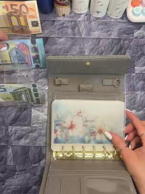 A close-up shot shows a person's hand with long, white-painted nails holding an open, gray wallet. The wallet is a bi-fold design with a gold clasp, and inside there's a decorative floral card. To the side, there are several Euro banknotes, including a 50 Euro and some smaller bills. The background is a textured, gray surface.