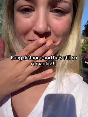 A woman with blonde hair and orange-tipped nails holds her hand over her mouth, looking emotional. Text overlaid says "Long distance and he's still so romantic!!!". The background is somewhat blurred, suggesting an outdoor setting.