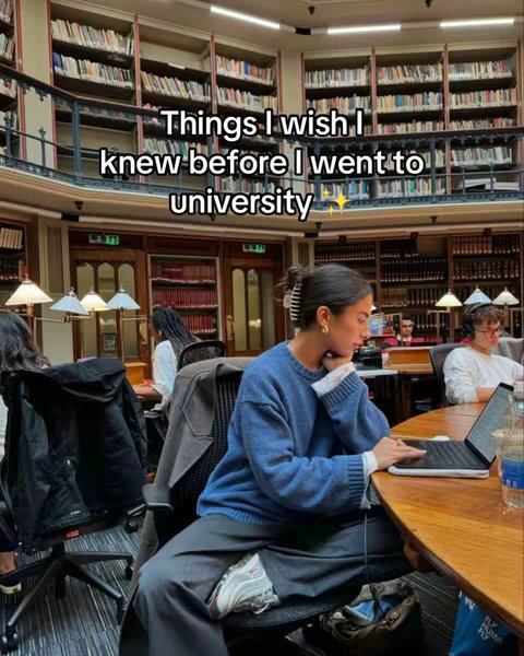 The image showcases a well-lit library interior with a large, circular table in the foreground. In the center, a person is seated at the table, engrossed in work on a laptop. The shelves are filled with many books and the overall composition highlights the ambiance of an academic library.