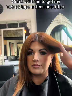 A woman with auburn hair is getting her hair styled, a person's hand is helping arrange her hair, with mirrors, a hair salon, and the woman's face taking up most of the image.
