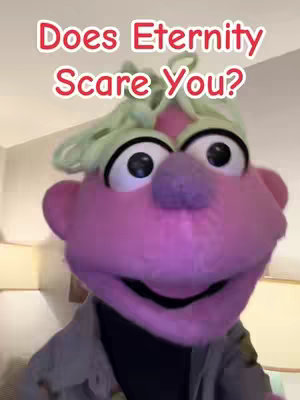 The cover image features a close-up shot of a puppet character. The puppet has a vibrant pink face, large black eyes, a purple nose, pink ears, and a large, smiling black mouth. It has light green, wavy hair on its head and is dressed in a gray jacket over a black shirt. The background is blurred, suggesting an indoor setting.