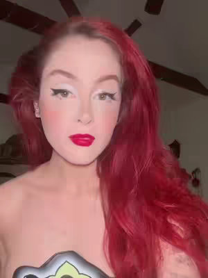 A close-up selfie of a person with bright red hair and bold makeup. They have a fair complexion, with defined eyebrows, winged eyeliner, and a bright red lipstick. Their cheeks have a rosy hue. The background is blurred, showing an indoor setting with ceiling fan blades visible. The person is facing the camera directly.