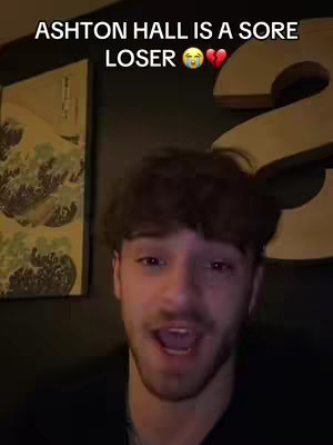 A man with short brown hair is in the foreground, looking at the camera and speaking. Overlaid at the top is white text that reads, "ASHTON HALL IS A SORE LOSER" with a crying emoji and a heart emoji. Behind him, there's a wall with art and a large letter "2". The lighting is dim.