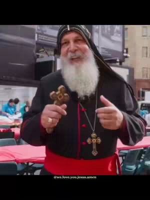 A man wearing religious attire, including a black robe and a white beard, holds a wooden cross in one hand and points to the viewer with the other in an outdoor setting. Background elements include table, other people, and tall buildings.