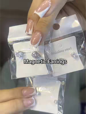 The image shows a close-up of a hand holding several small, clear plastic bags each containing a pair of stud earrings. The hand has well-manicured nails with a white french tip. Above the bags, is added text stating 'Magnetic Earrings.' The background is blurred.