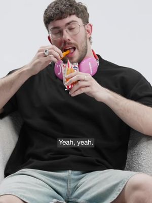 A man with glasses wearing a black t-shirt and light blue shorts is casually seated indoors, appearing to be relaxing while holding an open bag of chips, with headphones around his neck. A text overlay says 'Yeah, yeah.'