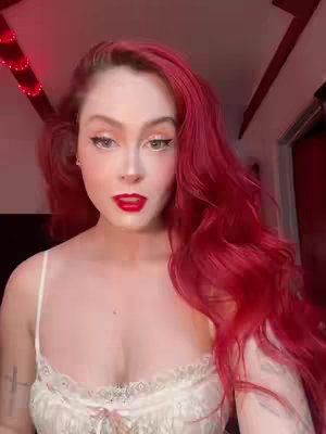 A close-up shot featuring a woman with vibrant red hair, red lipstick, and detailed makeup. She wears a white, lace-trimmed top, and the background has a red hue, likely from lighting.