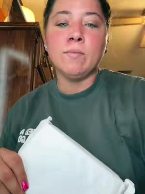 A woman with fair skin and light eyes looks directly at the camera. She has a slight smile and appears to be in a casual setting. She is holding a white package out in front. The background is slightly out of focus and suggests a warm, indoor environment.