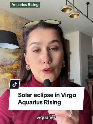 The cover image features a woman with dark hair and a maroon turtleneck sweater, speaking into a microphone. She is wearing glasses on top of her head and has decorative earrings. The background includes a textured wall, a black lamp, and a modern light fixture. Text overlays on the image read "Aquarius Rising" and "Solar eclipse in Virgo Aquarius Rising".