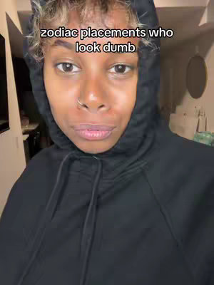 The image is a selfie-style shot featuring a person indoors. The person, a black woman, is positioned centrally in the frame with focus on the upper body and face. She's wearing a black hooded sweatshirt, and the hood is up, partially obscuring the top of her head and framing her face. Her hair is short, with some blonde highlights. The expression is neutral, with eyes looking directly at the camera. The background is blurred, suggesting an interior setting with a light-colored wall and a glimpse of some other objects, maybe furniture. The lighting is somewhat soft, as if the image was taken indoors. Overlay text reads: zodiac placements who look dumb.