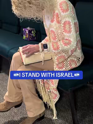 A woman in a patterned shawl sits on a teal seat, holding a book in her lap. A hook overlay stating "I STAND WITH ISRAEL" is on the bottom.
