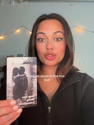 A woman with medium-length brown hair and makeup holds a tarot card in front of her. The card depicts two silhouetted figures embracing. A string of lights is visible in the background, above and behind the woman. Text is overlaid on the image, stating " you are about to find true love".