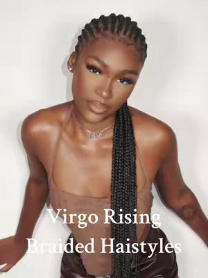 A woman with dark skin and braided hair is centered in the frame, looking towards the viewer. Her hair is styled in tight, small braids across the top of her head, with a long braid hanging down one side. She wears minimal makeup and a delicate necklace. The background is a plain white surface.