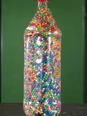 A clear plastic bottle shaped like a soda bottle is filled with a multitude of colorful spherical objects, primarily in shades of red, yellow, green, blue, and orange. The bottle has a red cap. The background is a plain, dark green surface, which provides a stark contrast to the brightly colored contents of the bottle. The overall image feels lively and visually engaging.