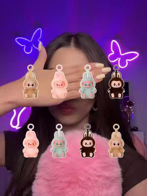 A selfie-style shot featuring a young woman with long, dark hair. She is seen from the chest up, with her face partially obscured by her left hand, revealing only her lips and part of her nose. The background is a deep purple, with two butterfly-shaped neon lights on either side of her head. Floating around her are eight cartoon rabbit keychains with different color schemes.