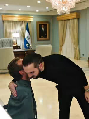 An image shows two men embracing indoors. The man on the right is older, wearing a black sweater, and has a full beard. He is embracing the younger man, whose back is to the camera, wearing a green jacket. The setting appears to be an office with a chandelier, drapes, a flag, and a painting on the wall.