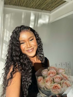 A woman with long, curly black hair smiles while holding a bouquet of pink roses. The background includes a wall and ceiling with a textured design.