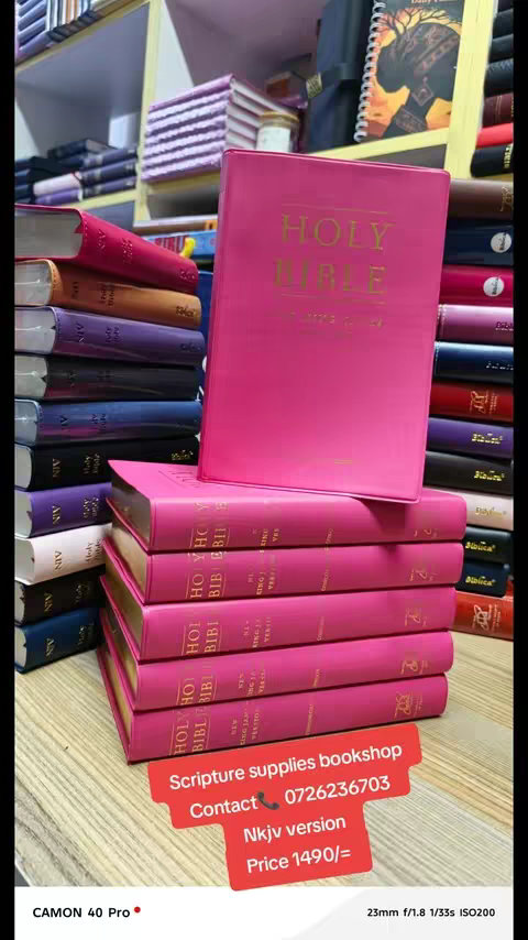 The image shows a display of various books, primarily Bibles, stacked and arranged to create a visually appealing bookstore setup. The dominant colors are pink and gold, with the Bibles showcasing pink covers and gold lettering. The Holy Bible text is visible on the front. They are placed on a wooden shelf with other books, some of whose spines are visible, adding depth and context to the scene. At the bottom, there is a red rectangular text box provides information, which reads 'Scripture supplies bookshop, Contact 📞 0726236703, Nkjv version, Price 1490/='.