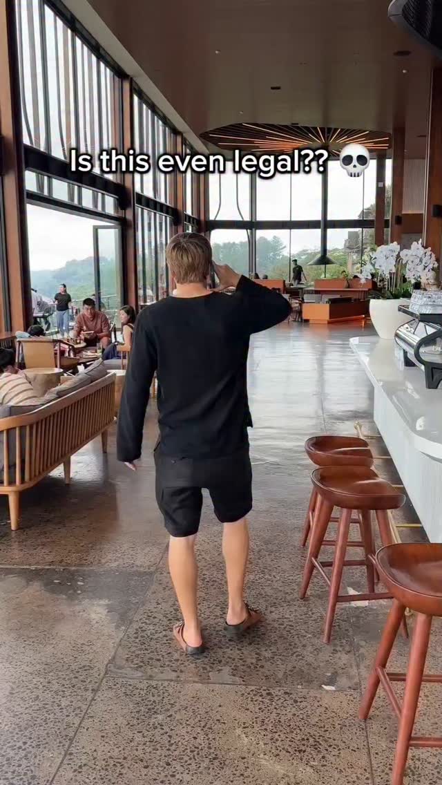 A person stands in a modern, open-plan space, likely a cafe or restaurant with large windows and a view of outdoor trees and landscape. The person is facing mostly away from the camera while holding their phone to their ear. Text overlayed on the image reads "Is this even legal??".