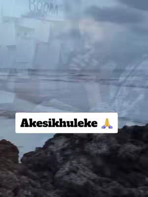The cover image displays a blurred, overcast seaside scene. In the foreground, dark rocks are visible. Above the rocks is a blurred view of what appears to be the ocean and cloudy skies. A white rectangle at the bottom of the image contains the text โAkesikhulekeโ in a bold, sans-serif font, with a small praying hands emoji on the right.