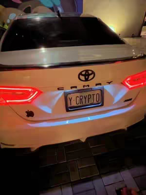 The cover image shows the rear of a white Toyota Camry. The license plate reads 'Y-CRYPTO.' An Apple logo sticker is visible on the left side of the trunk. The car is parked on a brick-patterned surface, with what appears to be exterior lighting. The taillights are illuminated.