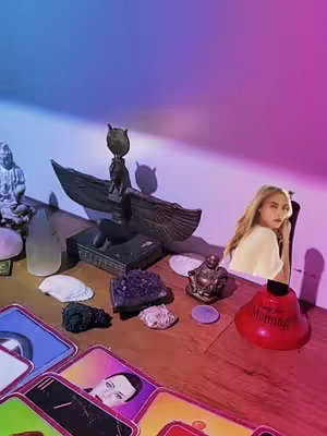 A still life arrangement, featuring a woman in a studio setting with mystical artifacts such as tarot cards, crystals, and a statue. The lighting shifts from blue to purple, creating a dreamlike ambiance.