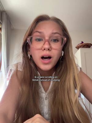 A young woman with long blonde hair and pink-framed glasses is the central figure. She is looking directly at the camera with an open mouth, possibly mid-sentence, and her eyes indicate a surprised or questioning expression. The background is softly blurred, with a glimpse of a window and a white cabinet visible. Text overlay adds information, and the overall lighting is bright.
