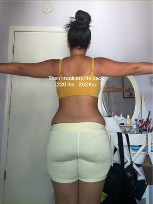 A woman stands with her back to the camera, arms outstretched horizontally. She wears a yellow sports bra and white shorts. Text overlay indicates weight loss. The background includes a door and a reflection in a mirror.