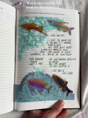 An open journal with a hand holding the page. The journal has handwritten text and stickers of fish on the page. The background is blurred, showing a soft, natural setting.