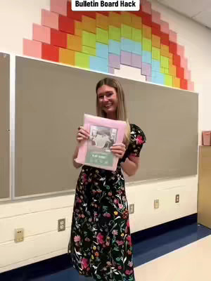 A woman stands in a classroom, holding a pink folder and smiling. She is wearing a long, dark dress with a floral pattern. Behind her is a bulletin board, some of which is plain but the top portion is adorned with a rainbow design made from colorful squares.