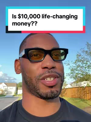 A man is captured in a selfie-style shot. He is wearing black-framed glasses with yellow-tinted lenses and a black t-shirt. He appears to be speaking toward the camera. Over his head is a banner with the words "Is $10,000 life-changing money??", set against a blue and red background. The background suggests an outdoor setting, with houses, trees, and a clear blue sky visible.