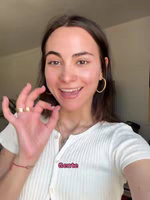 A young woman with dark hair and gold hoop earrings smiles and gives an 'okay' gesture. She wears a white, button-up shirt and a ring. A text overlay reads 'Gente' in a pink box.