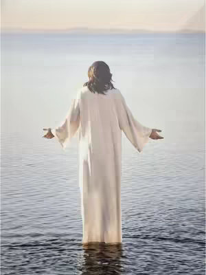 A serene image of a person standing in water, facing away from the viewer with arms outstretched. The person is wearing a long, white robe, and the background shows a calm body of water merging with a soft, overcast sky. The composition emphasizes peace and possibly spirituality.