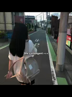 A person with long, dark hair walks down a street. They are wearing a white top, black shorts, and carrying a beige backpack. The street is lined with buildings and trees, with green painted lines along the sidewalks. Text overlay in white with black stroke reads, "She's intelligent >>>".