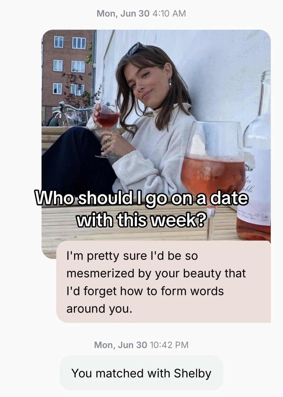 A woman is seated outdoors, holding a glass of red wine, with a second glass and wine bottle visible. The background shows a building and a pale wall.
