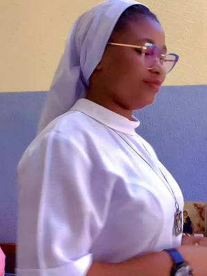 A woman wearing a white habit and glasses looks slightly right of the camera in this side profile shot. She has a soft expression and is wearing a small pendant necklace. The background shows a yellow and blue painted wall.