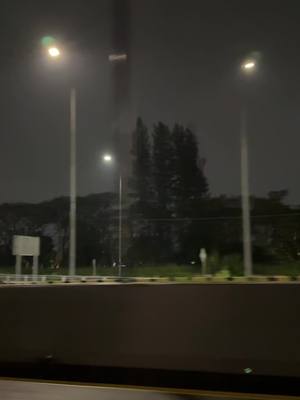 A nighttime outdoor scene with three streetlights prominently displayed. The image has a dark, moody aesthetic, with the sky appearing overcast or cloudy. The streetlights are evenly spaced, each with a bright, warm glow, illuminating the road and surrounding trees. The composition is balanced, with the lights creating a sense of depth and leading the viewer's eye into the background. The color palette is primarily dark, with the warm light of the streetlights contrasting against the cool tones of the night.